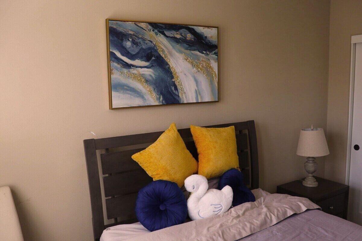 A bedroom with a bed, two pillows, a nightstand with a lamp, and a painting on the wall.