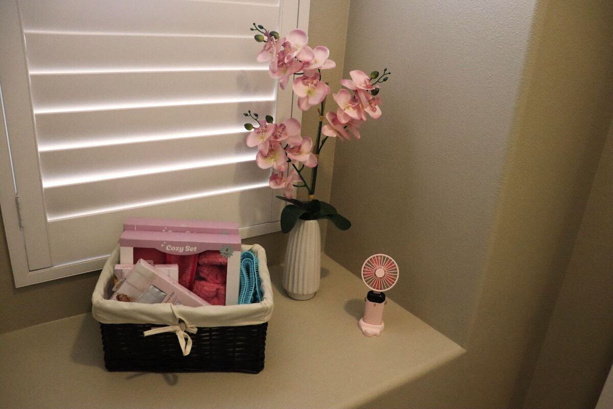 A wicker basket with a 'Cozy Set' box, a vase with pink flowers, and a small fan on a shelf.