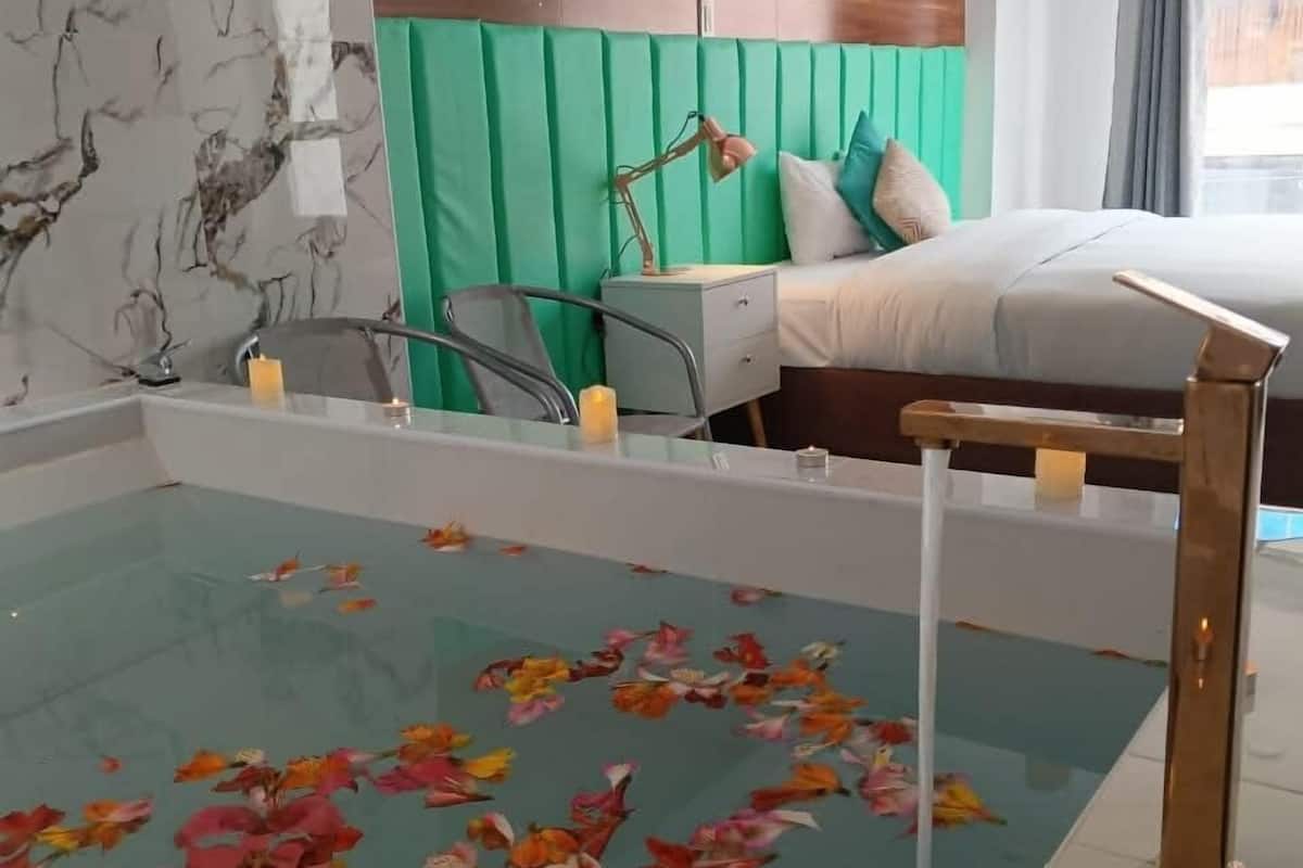 A modern hotel room with a large bed, a green padded headboard, a small desk with a lamp, and a glass-bottomed pool with floating flower petals.