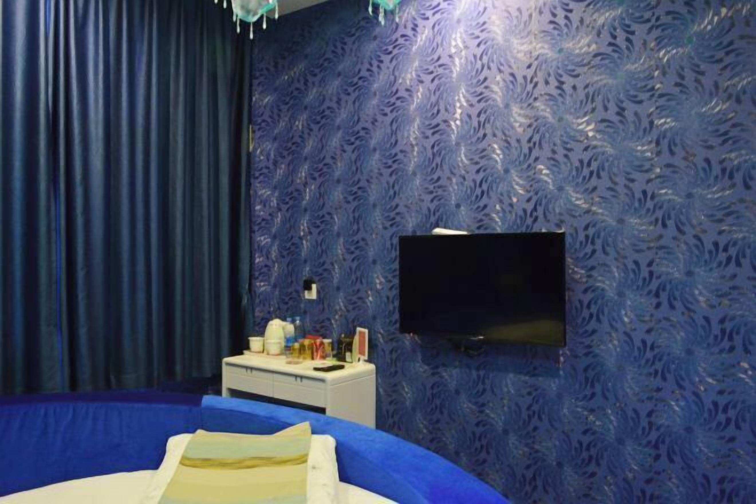 Une chambre avec un lit, un téléviseur à écran plat fixé au mur, une petite table de chevet avec une lampe et un rideau.