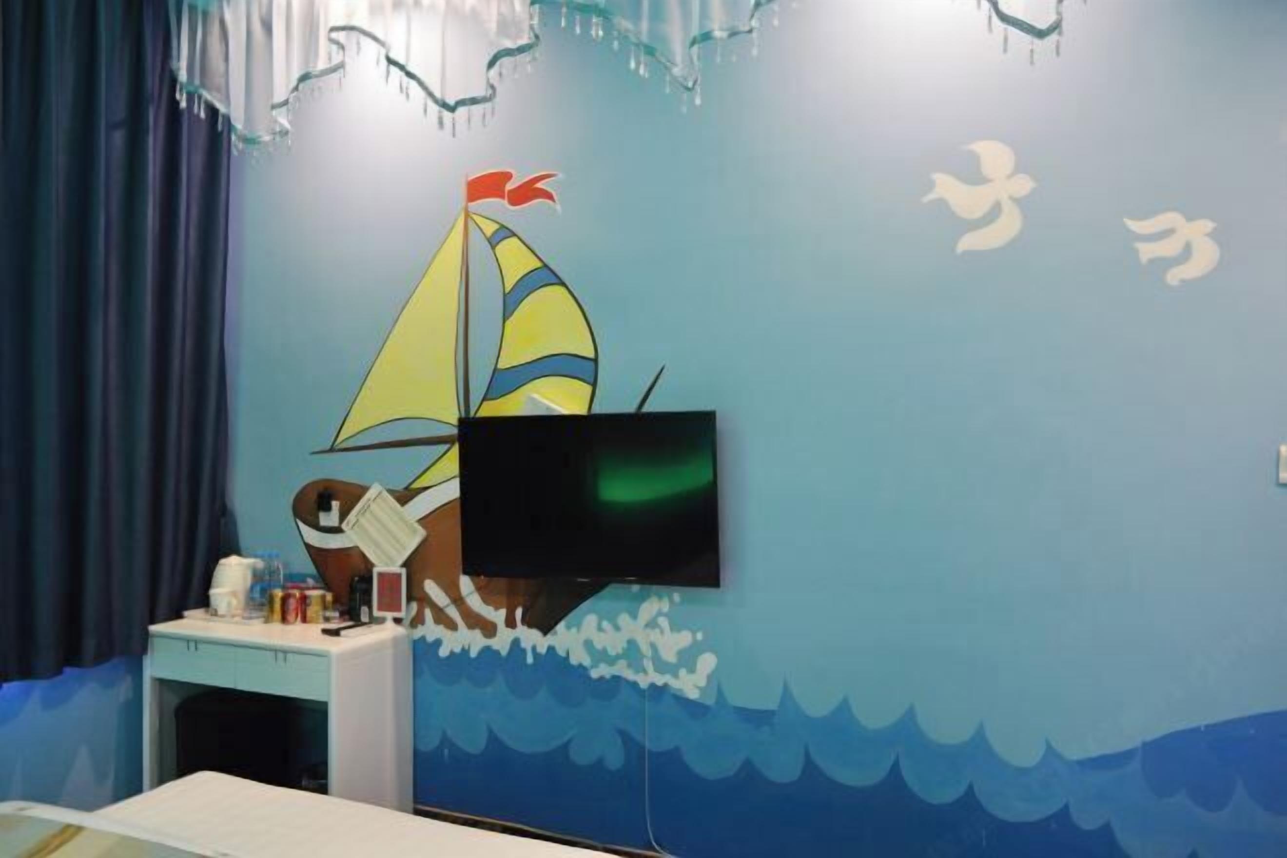 A room with a sailboat mural, a television mounted on the wall, and a desk with various items.