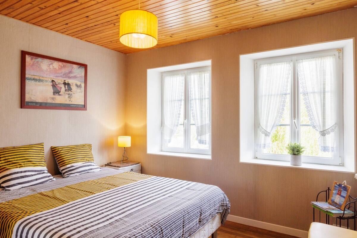A bedroom with a bed, two windows, a nightstand, a lamp, a painting, and a small table with a plant.