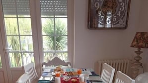 A dining table set with breakfast items, including bread, butter, and orange juice, in a room with a view of greenery through the windows. - AR VEIL (Plomeur)