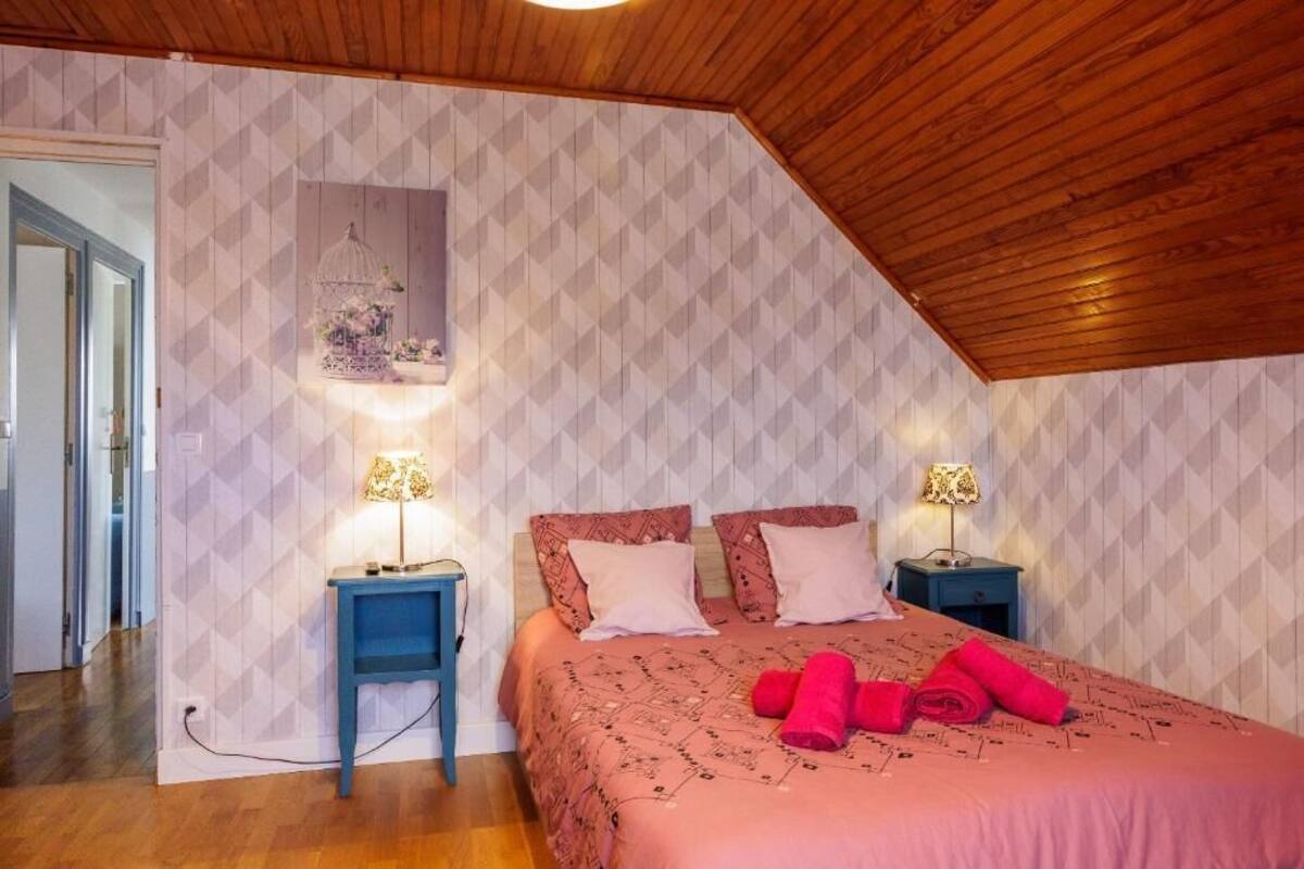A bedroom with a sloped ceiling, patterned wallpaper, two bedside tables with lamps, a bed with pink bedding and rolled towels, and a framed picture on the wall.