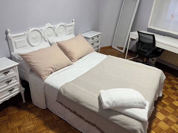 Una cama bien hecha con cabecera blanca, dos almohadas color beige y una colcha blanca. Hay dos mesitas de noche blancas con cajones a ambos lados de la cama.