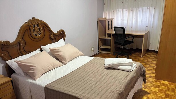Un dormitorio con cabecera de madera, cama con ropa de cama color crema, escritorio con silla y ventana con cortinas.