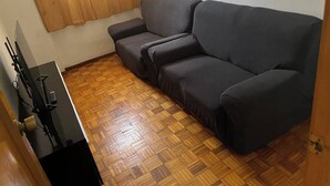 Una habitación con un sofá gris oscuro, suelo de madera en diseño de espiga y una pequeña mesa negra con dispositivos electrónicos.