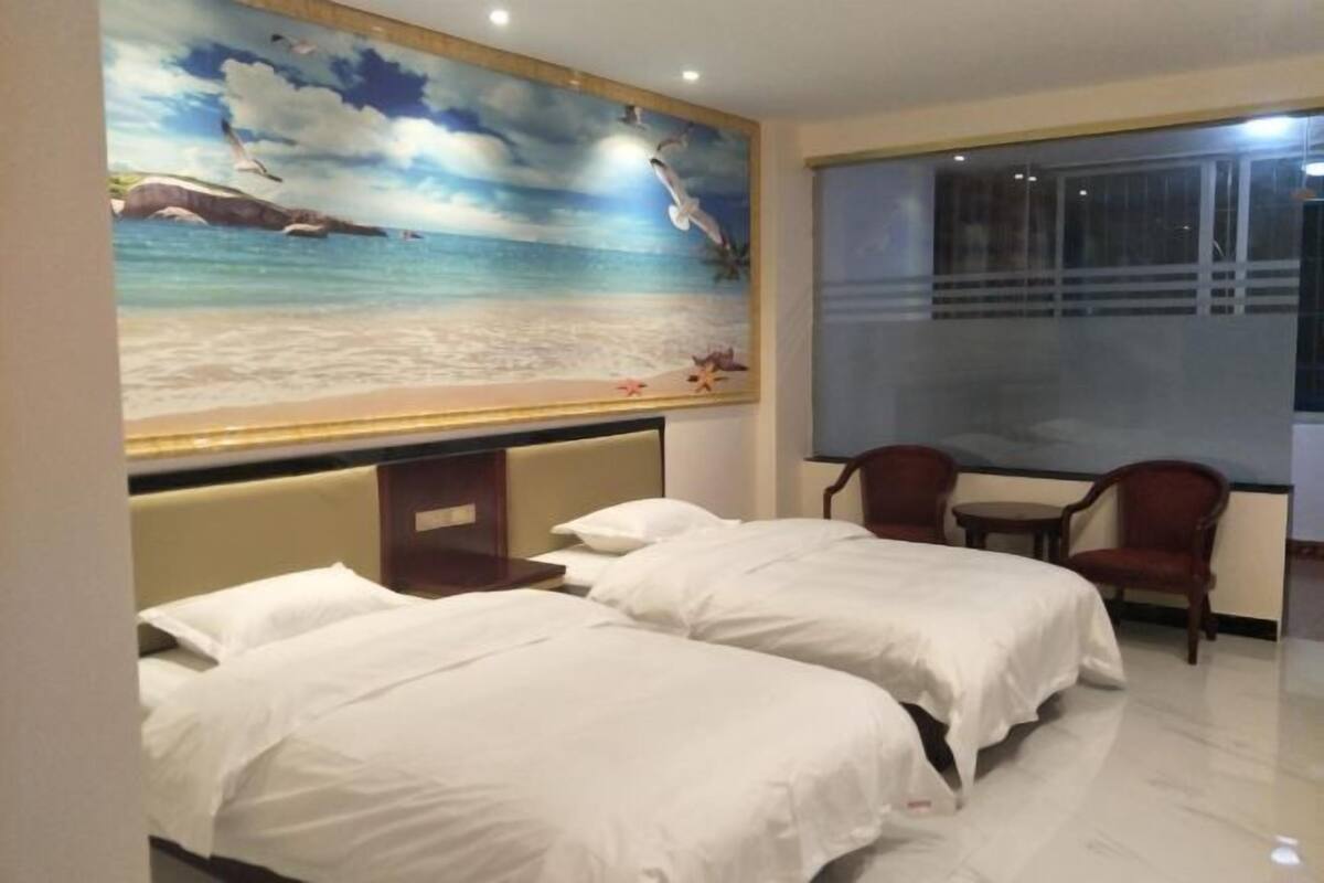 Habitación de hotel con dos camas, un mural grande de playa, una ventana con cortinas y una pequeña mesa con sillas.