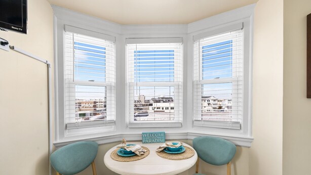 Une chambre d’angle avec un bow-window, une petite table avec deux chaises et une pancarte indiquant : « Bienvenue dans notre petit coin de paradis ».