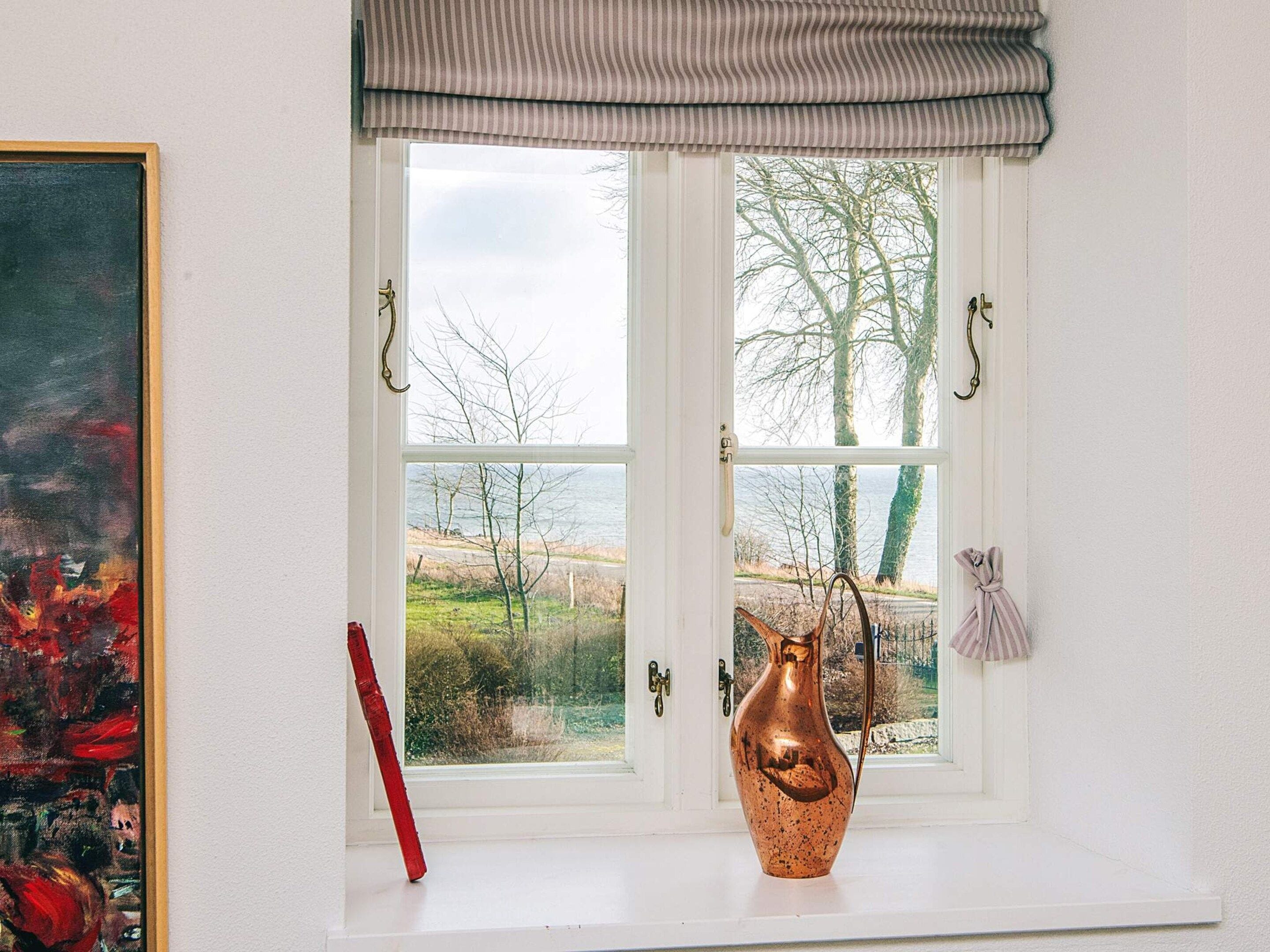 Una ventana con vista a árboles y un cuerpo de agua, un florero de cobre en el alféizar, y un cuadro en la pared.