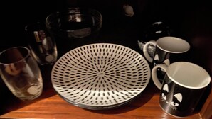 Ein Holztisch mit einer weißen, gelochten Schüssel, einer schwarzen Tasse mit Katzenmotiv und mehreren klaren Gläsern.