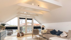 Un appartement moderne de style « loft », avec un plafond en pente, un ensemble de canapés, une table basse et une vue sur le paysage urbain grâce à de grandes fenêtres.