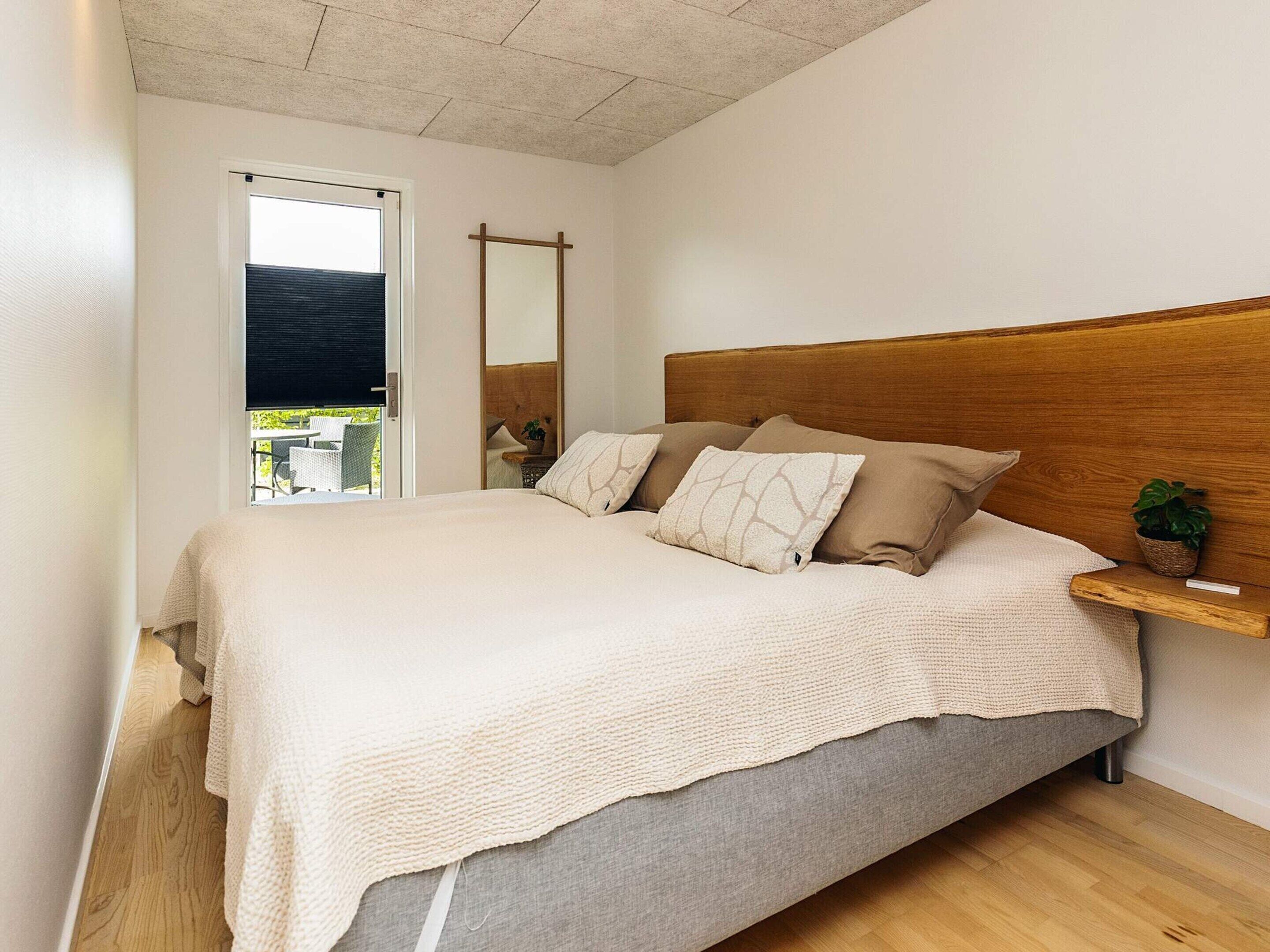 A bedroom with a bed, a wooden headboard, a bedside table with a plant, and a view through a window.