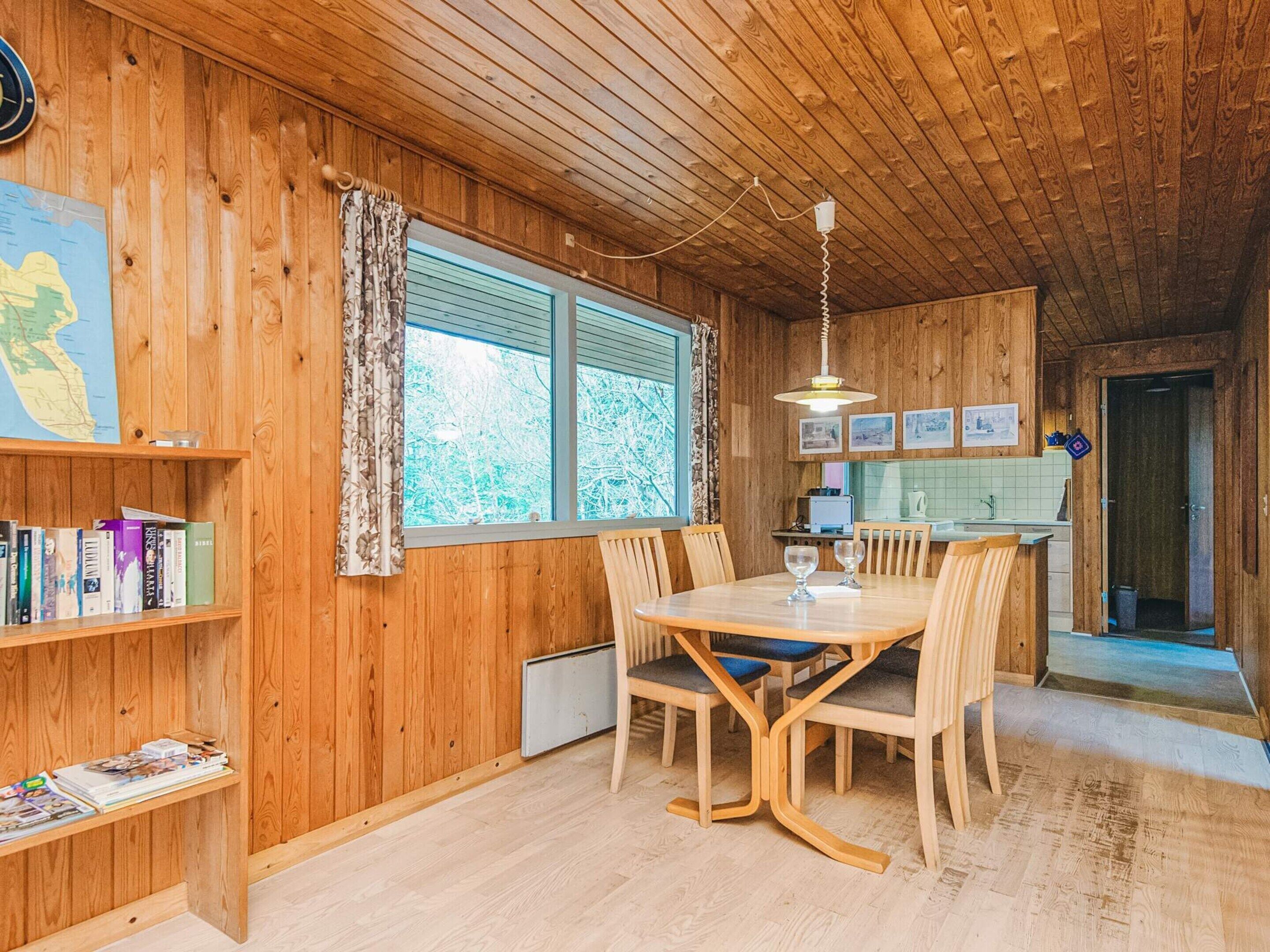 A wooden cabin interior with a dining table, chairs, a bookshelf, and a window with curtains.
