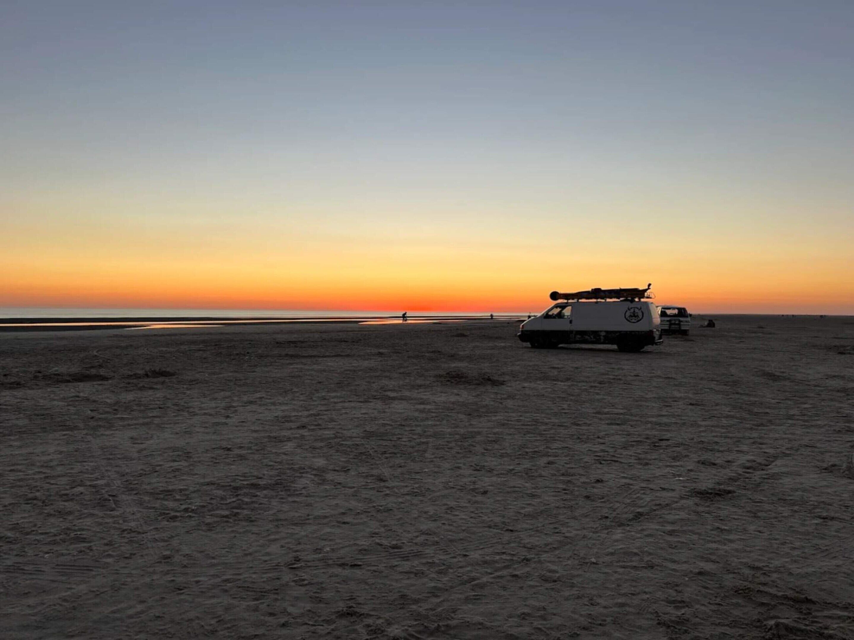 Een strandscène met een wit voertuig geparkeerd in het zand, een heldere lucht en een ondergaande zon op de achtergrond.