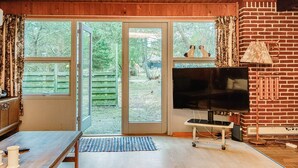 Une pièce chaleureuse avec un plafond en bois, un téléviseur posé sur un meuble, une table à manger et une grande fenêtre donnant sur un jardin.