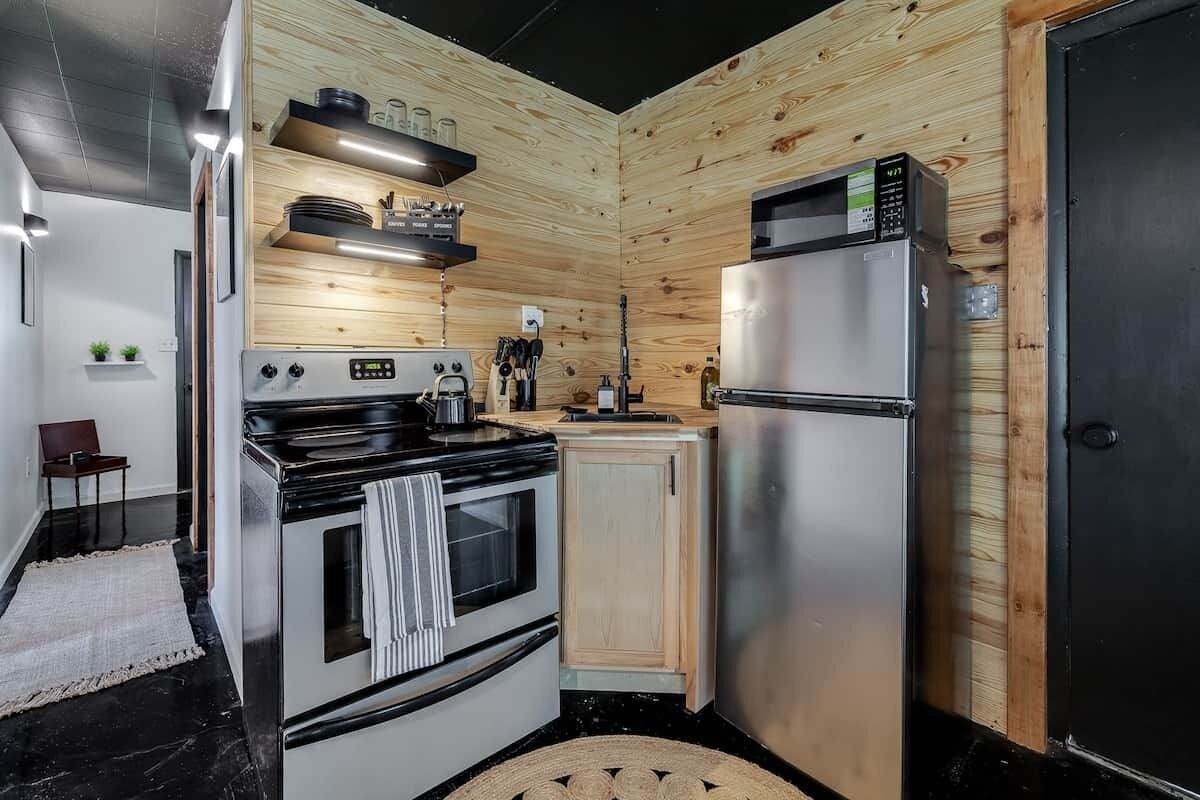 A compact kitchen with a stove, oven, microwave, and refrigerator, wooden panel walls, and a small countertop with utensils.