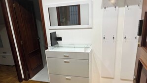 Una habitación con piso de madera, un armario blanco con cajones, un escritorio con cubierta de vidrio, un espejo y un guardarropa con ganchos.