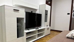 Una habitación con una televisión sobre un mueble blanco, una cama con un cojín estampado y suelo de madera.