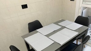 Una mesa rectangular con cuatro sillas, cada una con asiento negro y base de madera. La mesa tiene una superficie gris y está colocada frente a una pared con azulejos blancos.