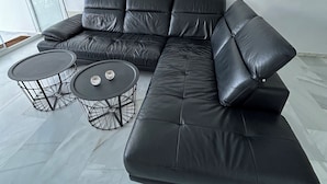 Un salon moderne avec un canapé d’angle en cuir noir, deux tables basses à structure métallique noire et une grande fenêtre qui laisse entrer la lumière naturelle.