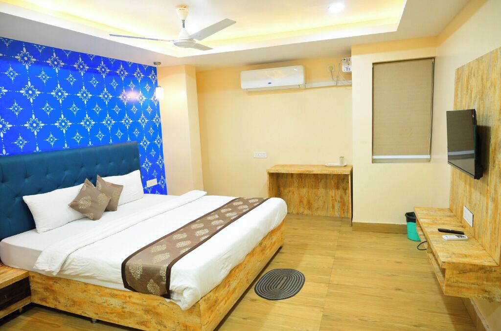 A bedroom with a large bed, a ceiling fan, an air conditioning unit, a TV, and a small wooden desk.