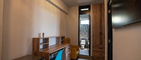 A compact bathroom with a wooden vanity, a small desk, a blue chair, and a toilet visible through an open door.