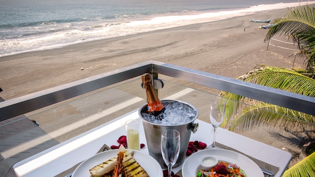 Ein Tisch mit zwei Teller Essen, einer Champagnerflasche und einem Weinglas, mit Blick auf einen Sonnenuntergang von einem Balkon über dem Strand.