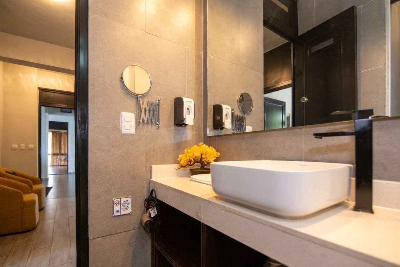 A modern bathroom with a large mirror, a white sink, and a small yellow flower arrangement.