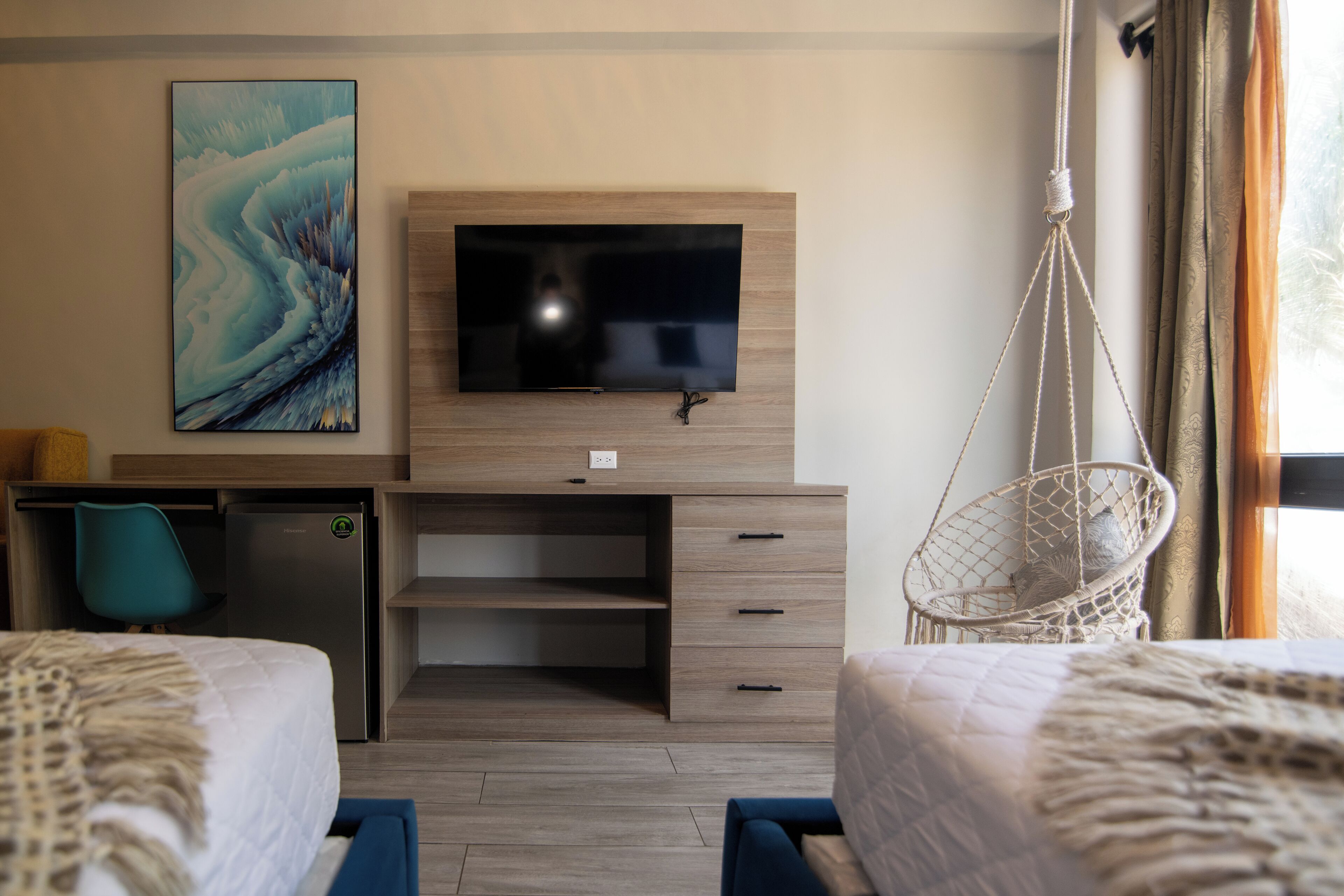 A hotel room with a flat-screen TV mounted on a wooden wall, a desk with a chair, a hanging chair, and two beds with textured bedding.