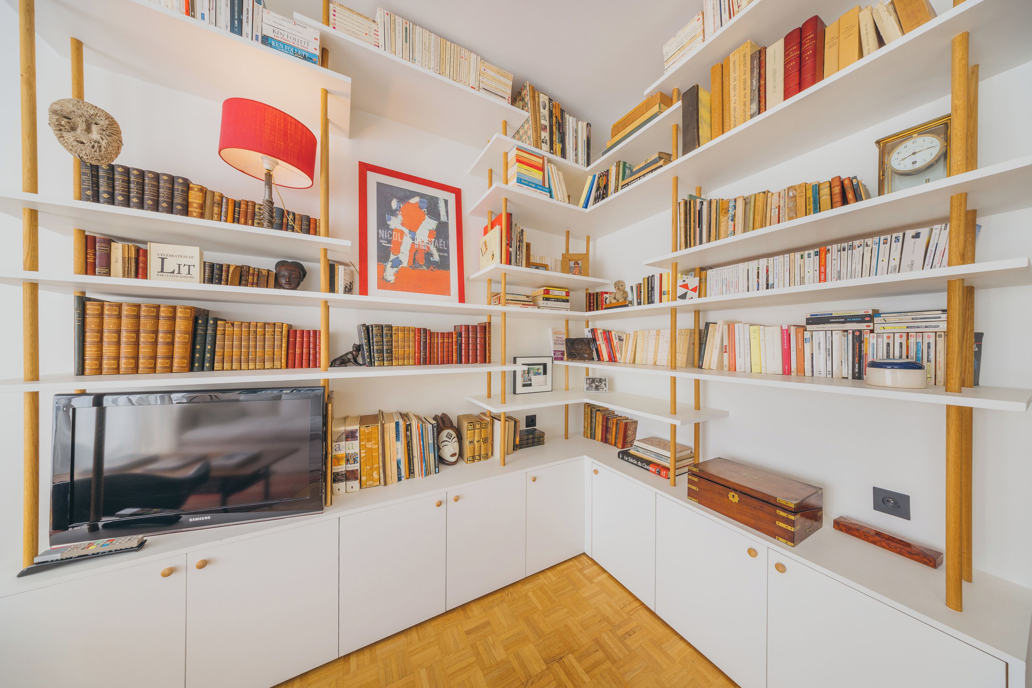 Una stanza ordinata con mensole bianche piene di libri, una lampada rossa e un televisore su un mobile.