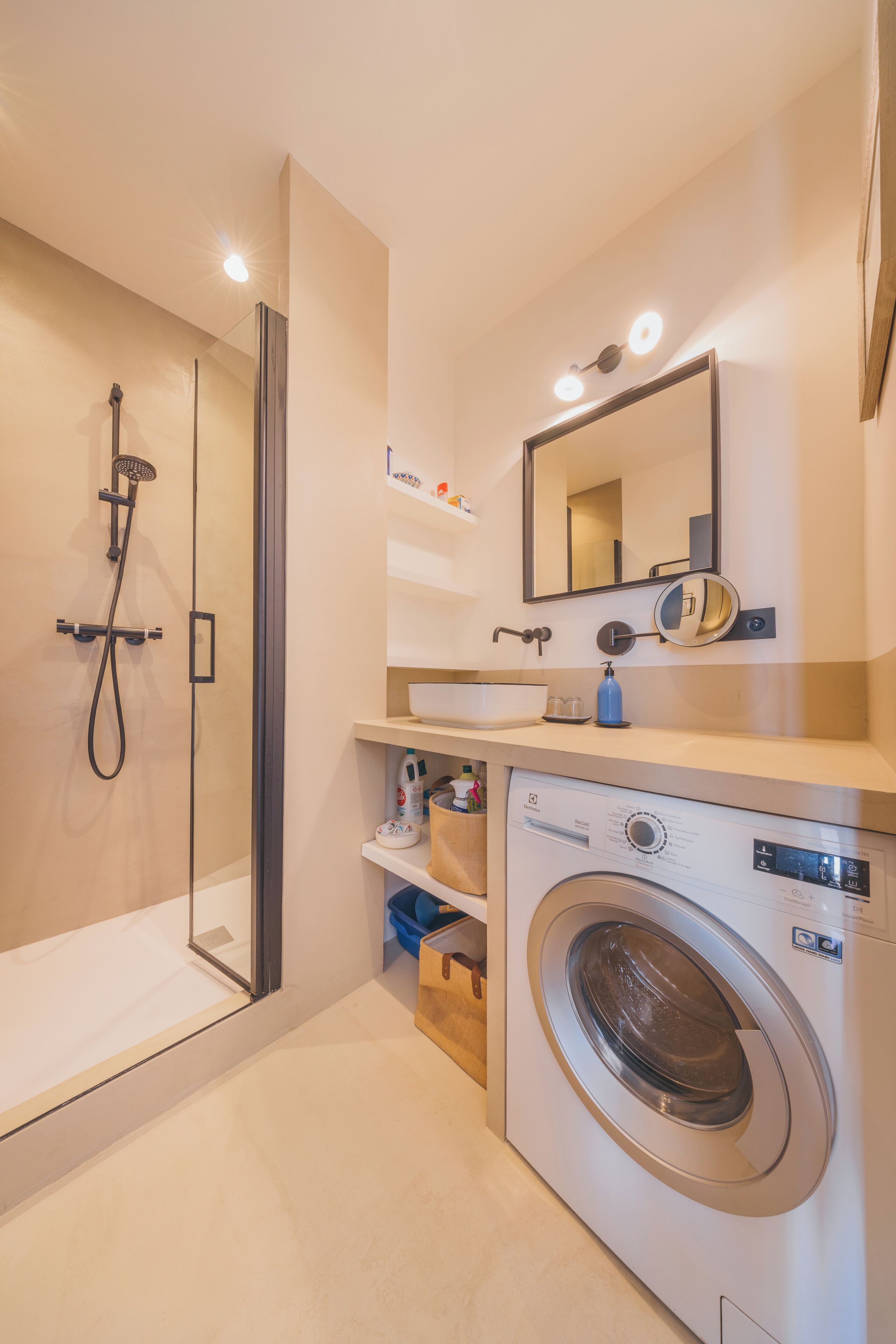 A modern bathroom with a glass-enclosed shower, a built-in sink, a mirror, and a front-loading washing machine.