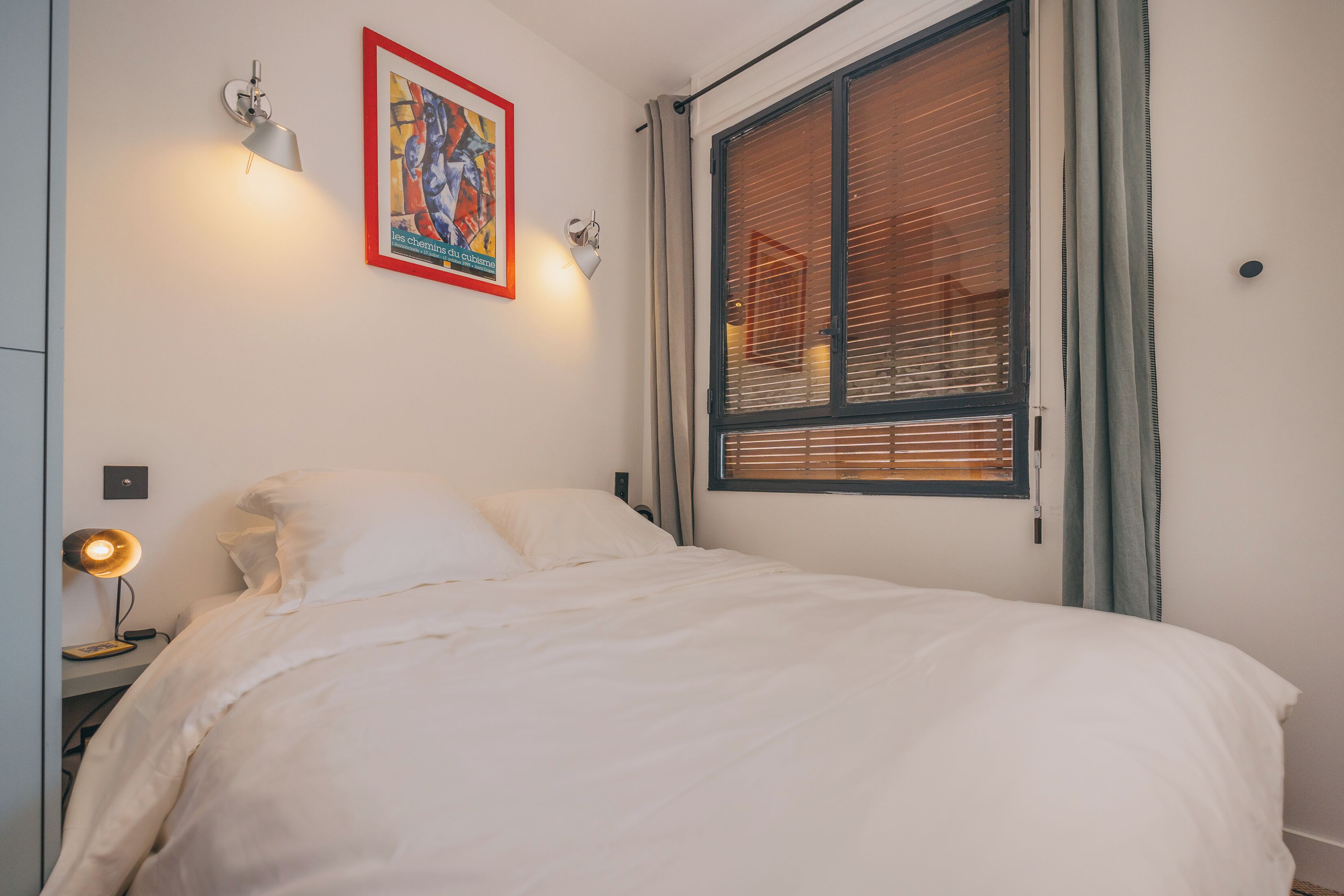 Un lit bien fait, avec une literie blanche, une lampe fixée au mur et une fenêtre équipée de stores.