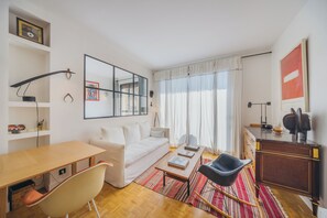 Une chambre d’hôtel moderne avec un bureau en bois, un canapé blanc, une chaise noire, un tapis rayé rouge et blanc, et une grande fenêtre avec des rideaux légers.