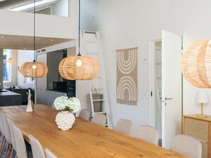 Une cuisine moderne avec une table à manger en bois, des lampes suspendues en osier et un mur blanc orné d’une tapisserie aux motifs géométriques.