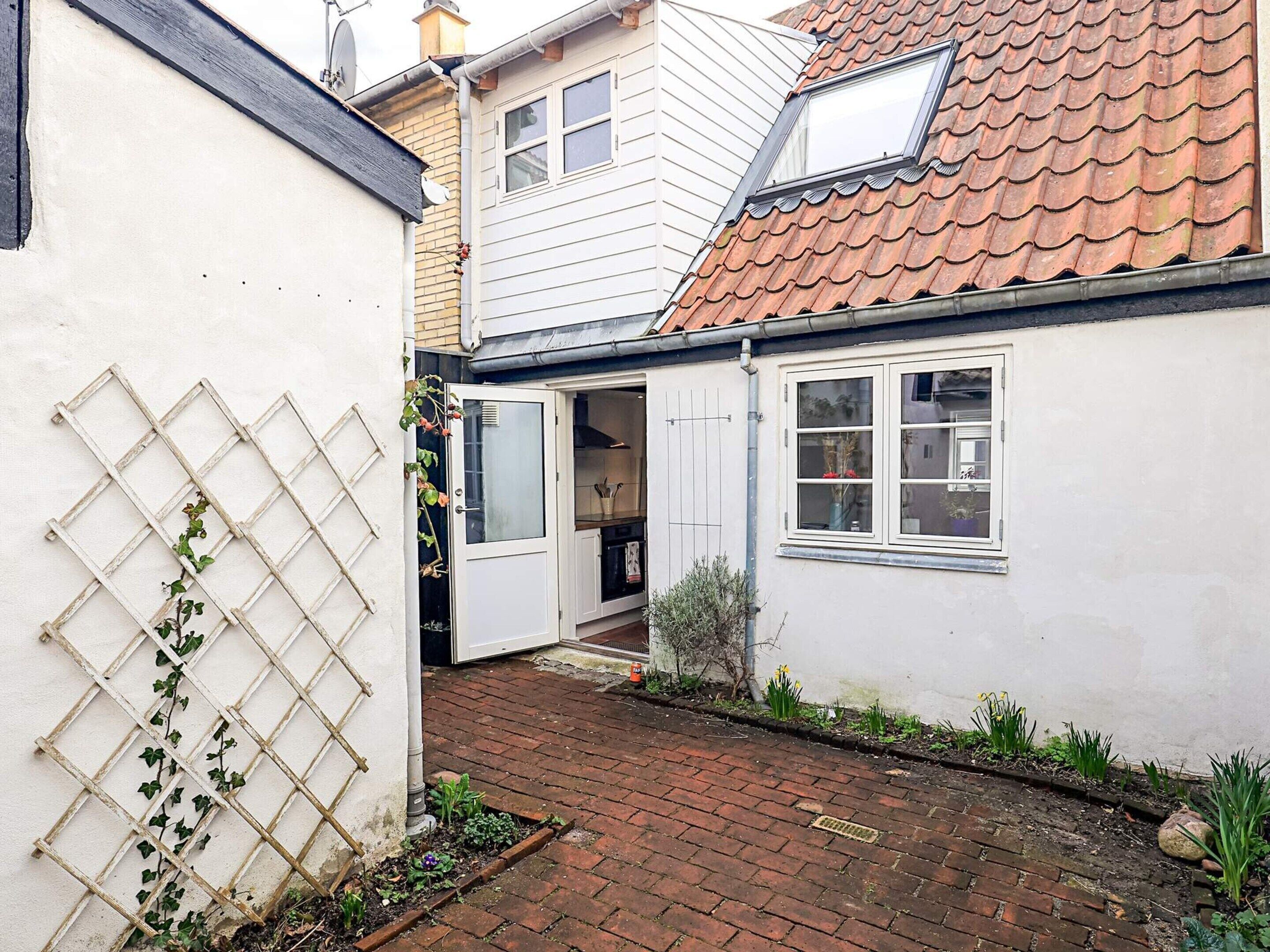 Un étroit sentier pavé de briques mène à une maison blanche avec un toit en tuiles et un petit jardin.