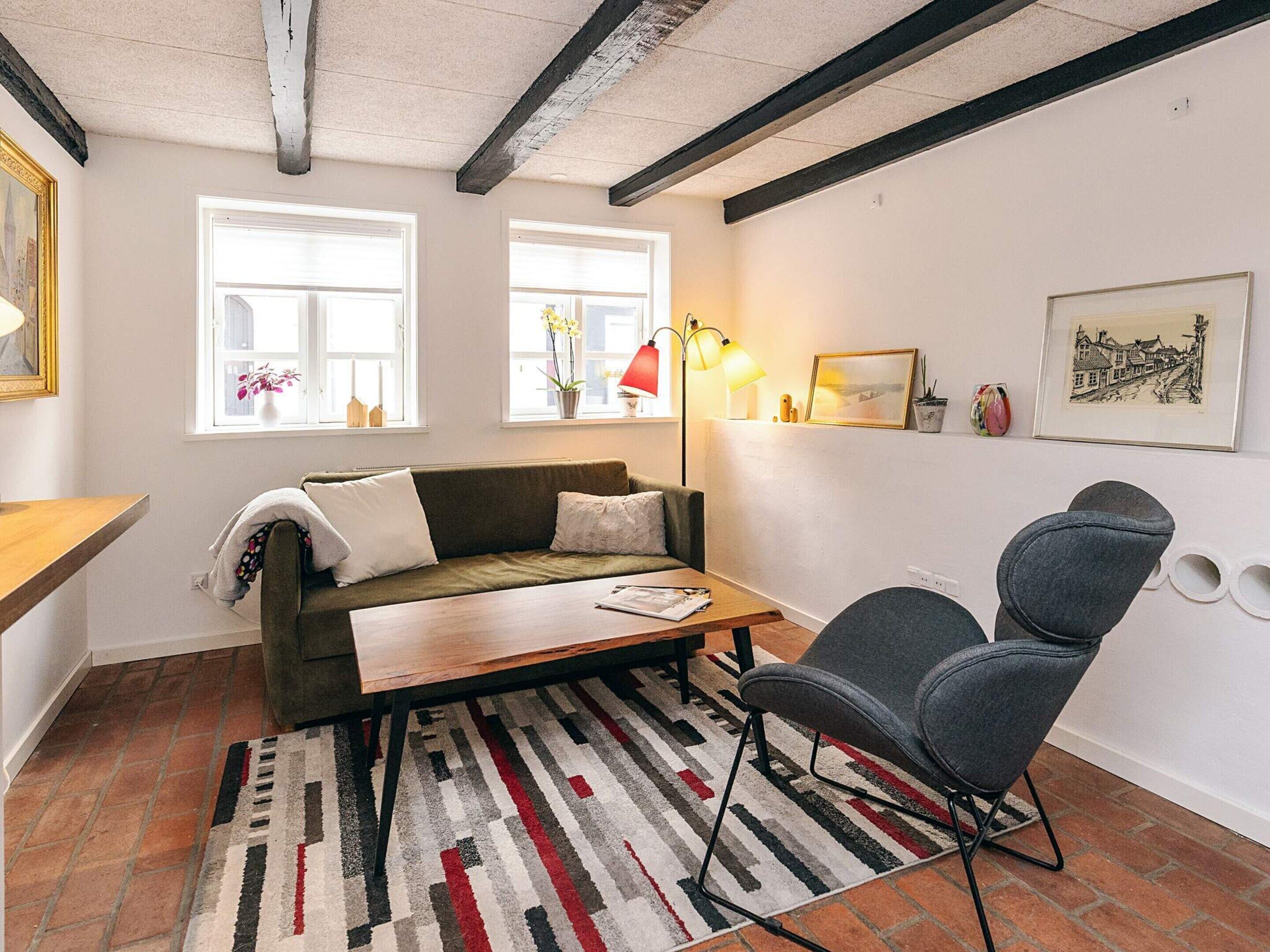 Un saló acollidor amb un sofà, una taula de centre i una cadira. Hi ha dues finestres amb cortines, una làmpada de peu i un quadre encimbellat a la paret.