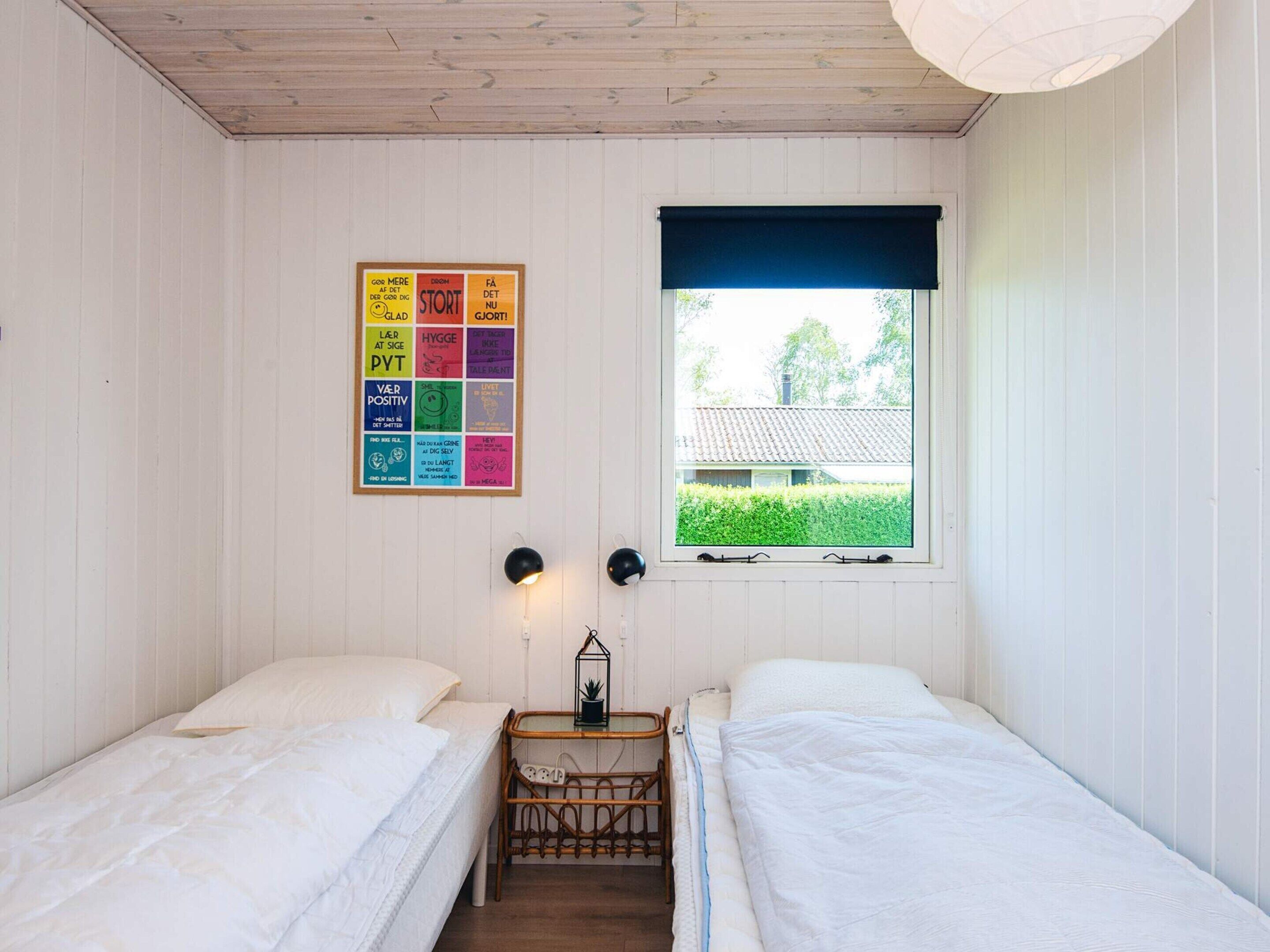 Una habitación con dos camas, una ventana con persiana azul, una lámpara de pared y un tablón de anuncios con notas de colores.