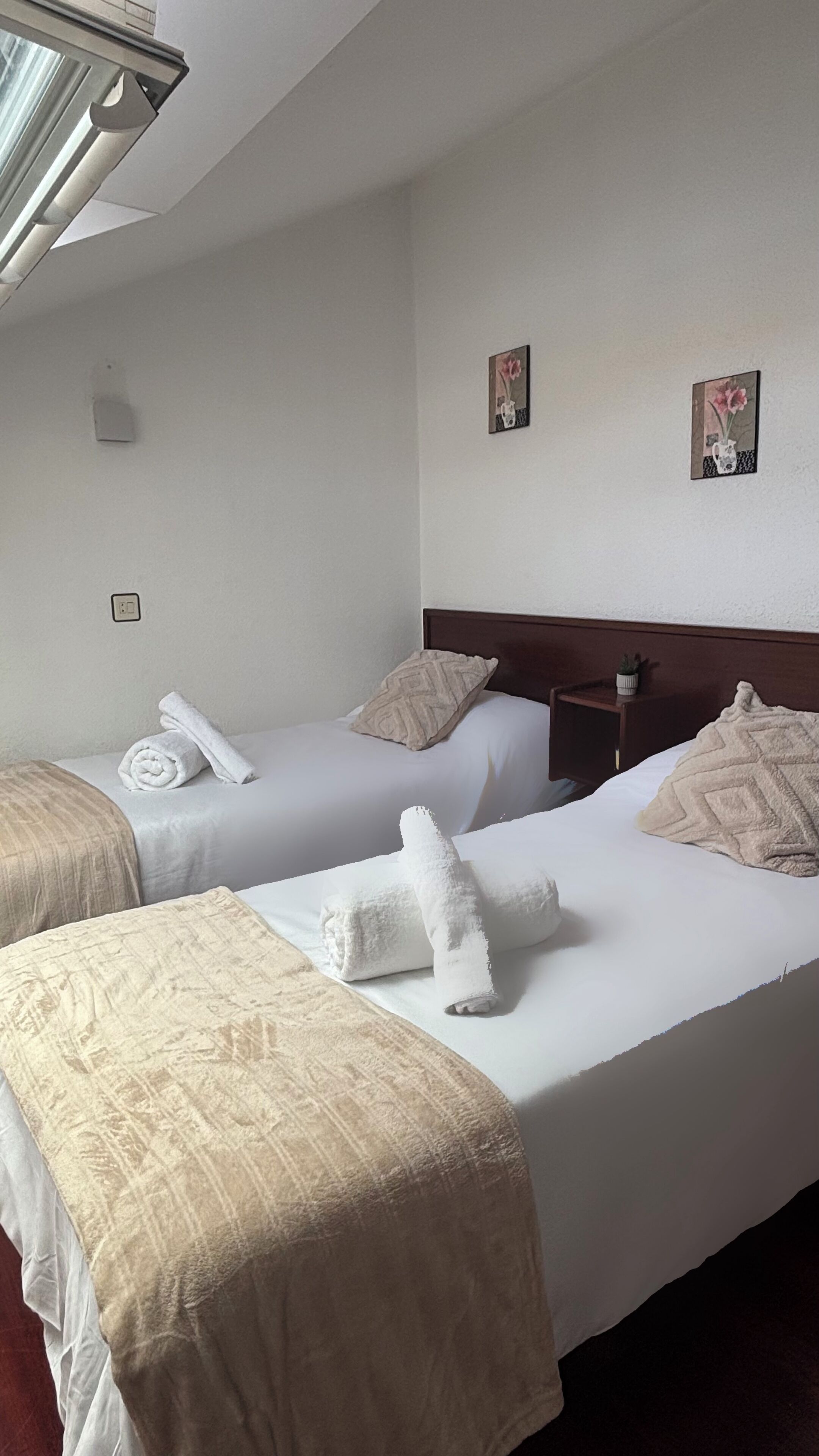 Habitación de hotel con dos camas, cada una con una colcha blanca y una manta color beige. Hay dos mesitas de noche fijadas a la pared, con lámparas, una ventana con persianas, y dos pequeños cuadros enmarcados en la pared.
