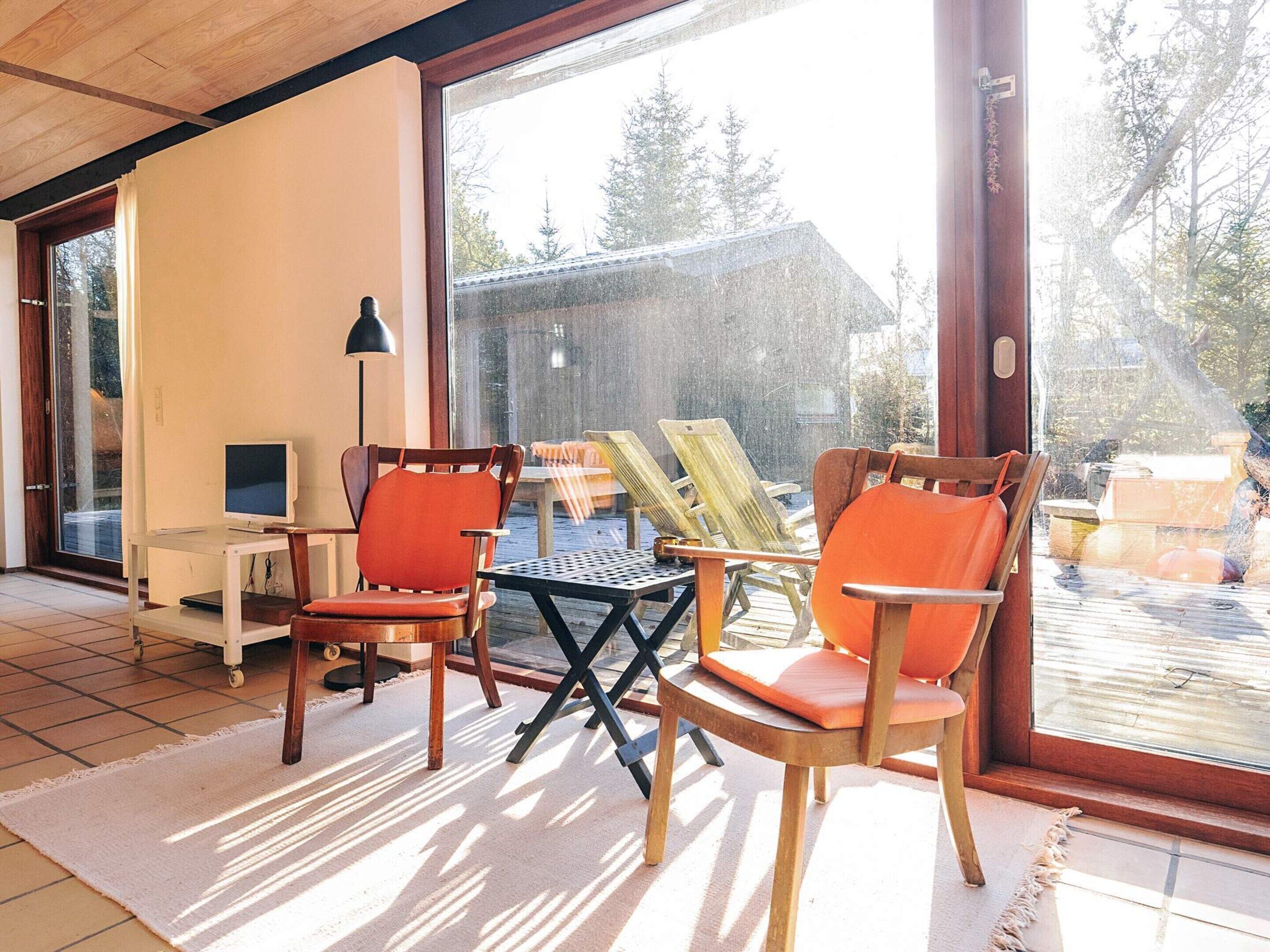 Um quarto iluminado pelo sol com uma grande porta de vidro deslizante que dá para um deck. No interior, há cadeiras de madeira com almofadas laranja, uma mesa pequena e um abajur.