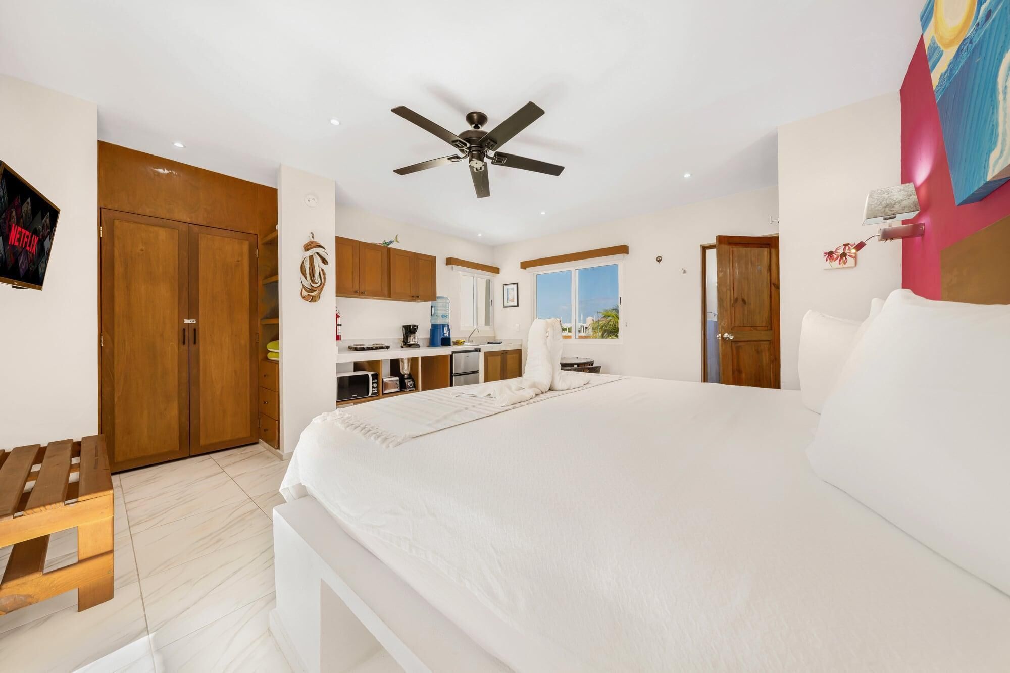 A bedroom with a large bed, a ceiling fan, wooden cabinets, a small kitchenette, and a TV on the wall.