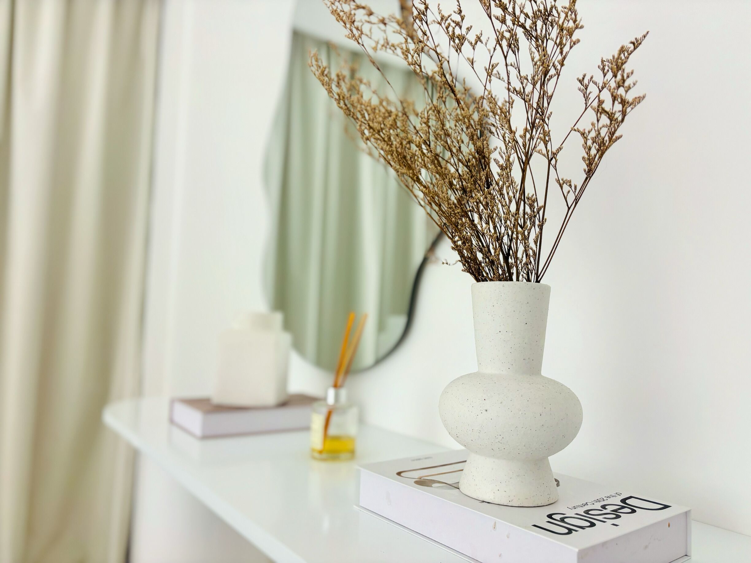 A white vase with dried plants on a white shelf.