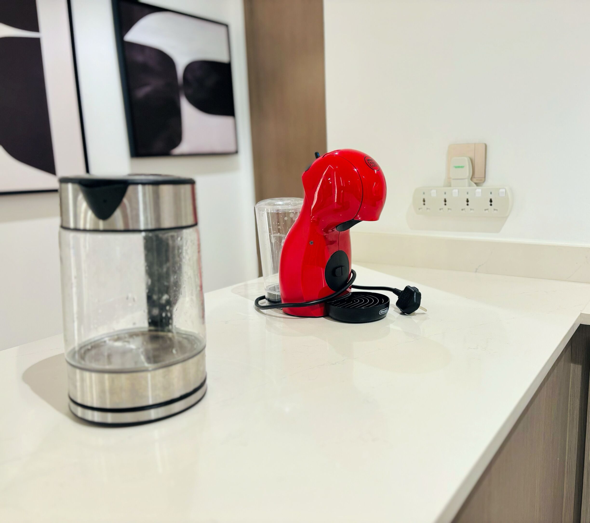 Un secador rojo sobre una encimera blanca, un vaso de agua transparente y una tetera de acero inoxidable.