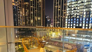 A modern hotel room with a large window overlooking a cityscape at night. The room features a glass table with two wicker chairs and a vase with flowers.