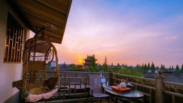 A balcony with a wicker chair, a table, and a view of a traditional building at sunset. - Chang'an Hotel (Zhujiajiao Ancient Town Branch, Qingpu, Shanghai) (Shanghai)