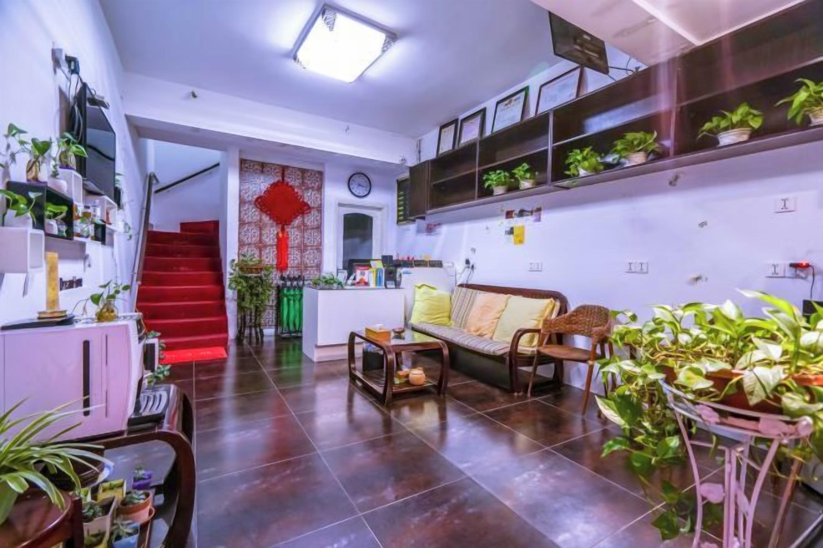 A living room with a sofa, a coffee table, and a dining table with chairs. There are potted plants and a staircase leading to an upper level.