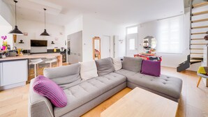 A modern living room with a grey sectional sofa, a wooden coffee table, and a kitchen area with a dining table and bar stools.