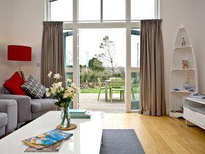 A modern living room with a sofa, a red lamp, a vase with flowers, a coffee table with magazines, and a view through a glass door to an outdoor area with a chair and greenery.