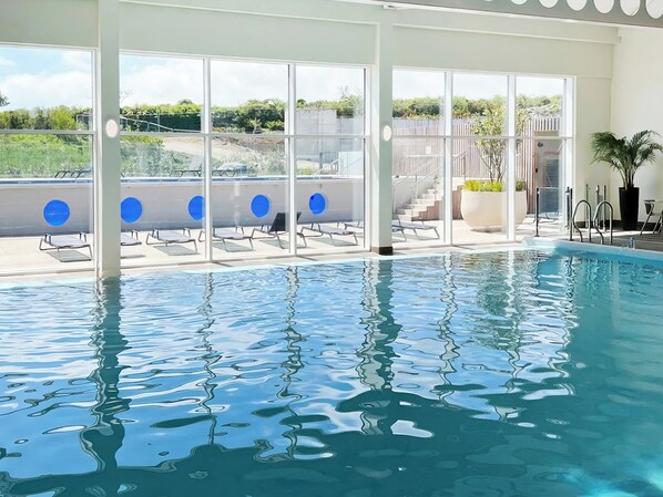 A swimming pool with clear water, sunlit interior, and a view of greenery outside.