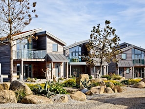 A modern multi-story house with a balcony, large windows, and a landscaped garden with rocks and plants.